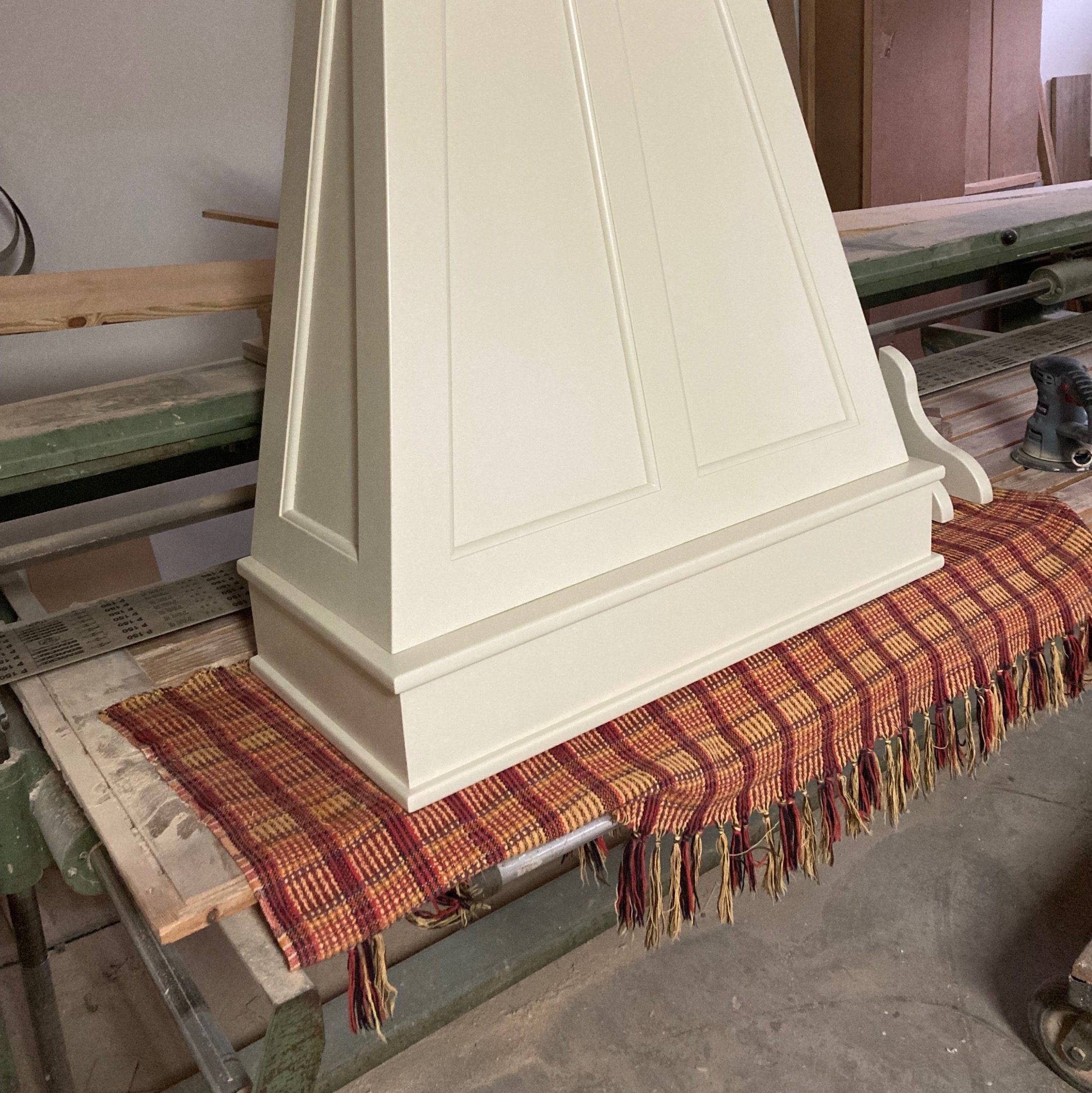 Biały, drewniany okap kuchenny w stylu klasycznym, ustawiony na stole warsztatowym przykrytym wzorzystą tkaniną z frędzlami, obok szlifierka elektryczna.