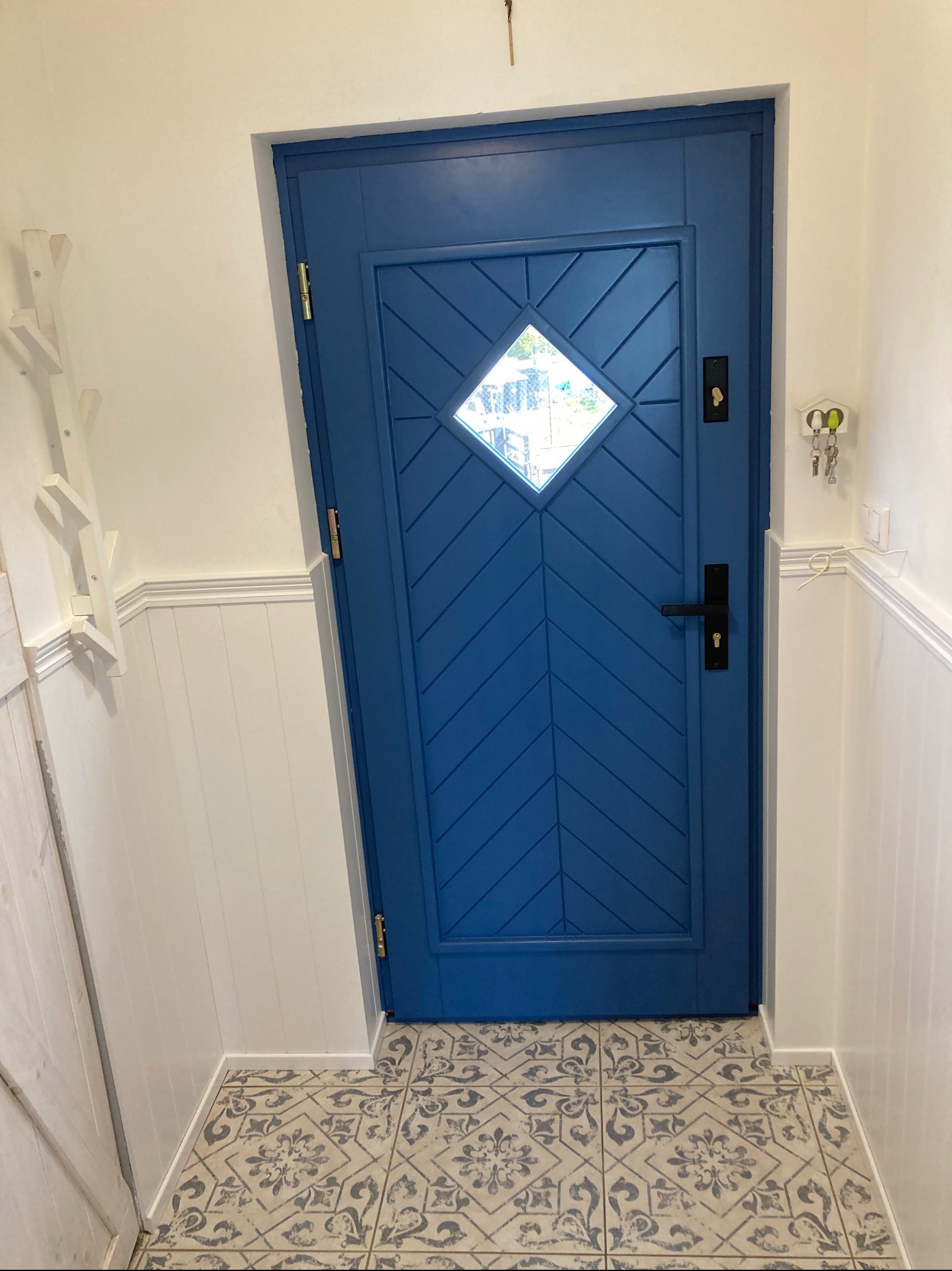 Niebieskie drzwi wejściowe z romboidalnym okienkiem, zdobione wzorem w jodełkę, zamontowane w białym przedpokoju z dekoracyjną boazerią i wzorzystą podłogą.