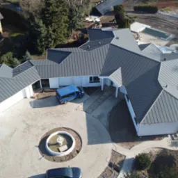 Widok z drona na rozległy, parterowy dom z szarą, metalową dachówką, fontanną na podjeździe z kostki brukowej i niebieskim busem zaparkowanym obok.