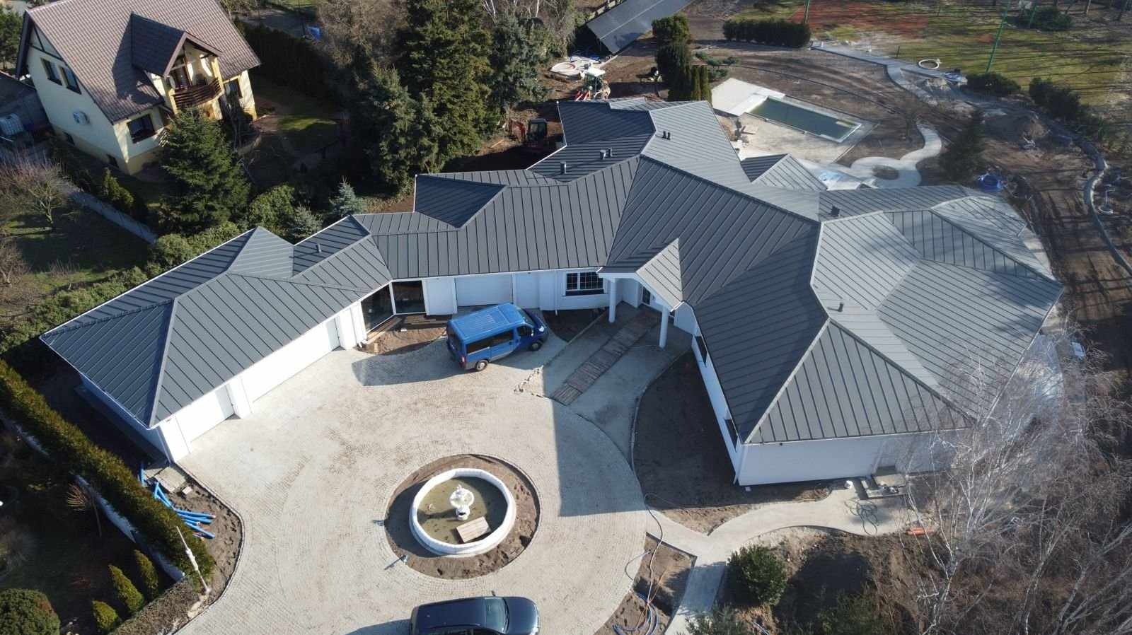 Widok z drona na rozległy, parterowy dom z szarą, metalową dachówką, fontanną na podjeździe z kostki brukowej i niebieskim busem zaparkowanym obok.