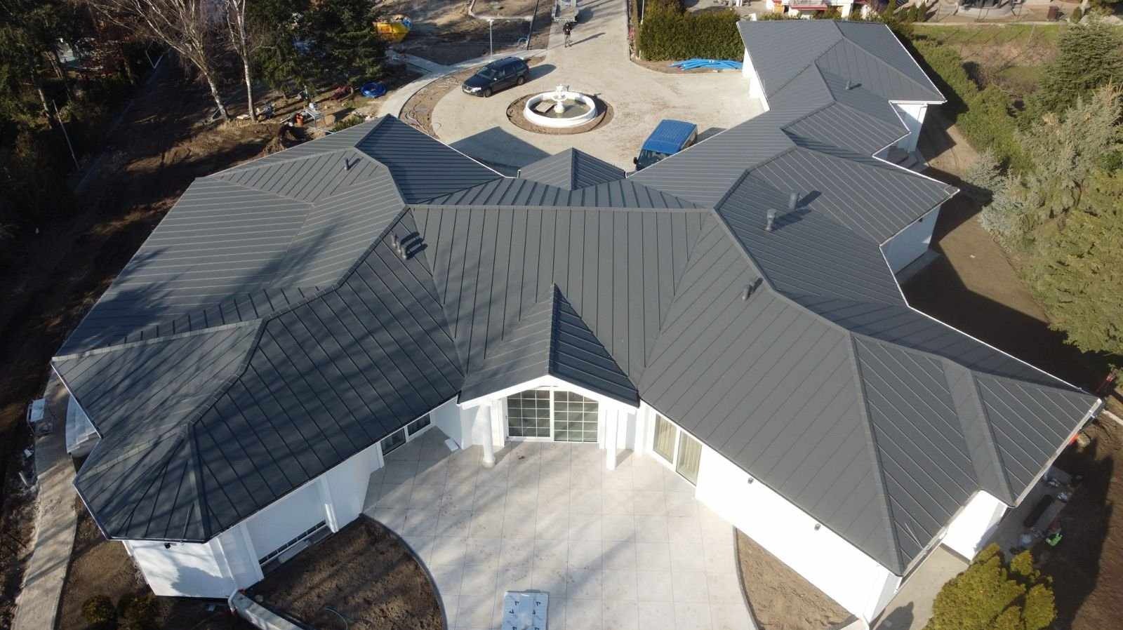 Widok z lotu ptaka na duży, biały dom z szarym, metalowym dachem wielospadowym. Na podjeździe fontanna i zaparkowane samochody.