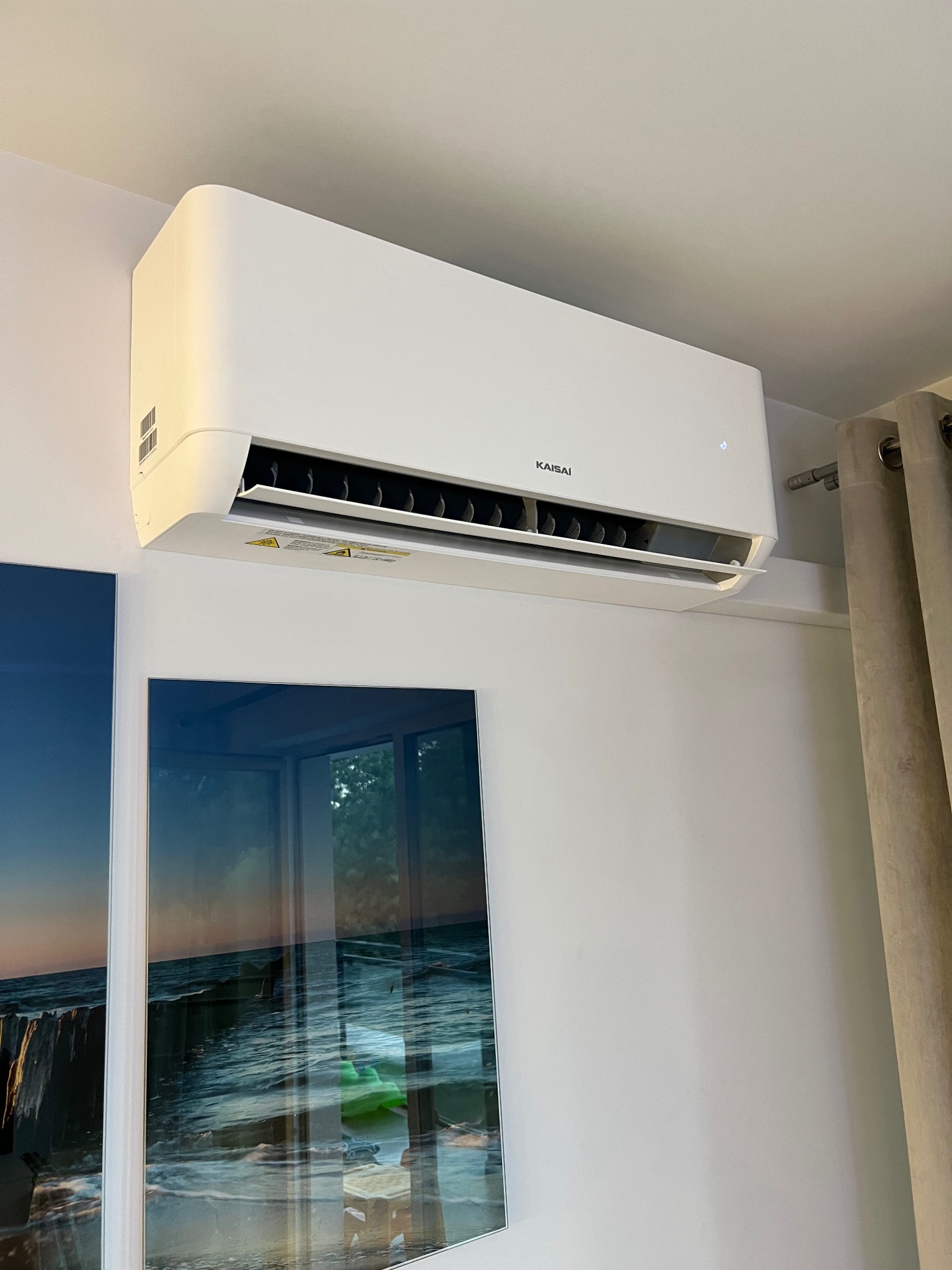Biały klimatyzator Kaisai na ścianie, pod sufitem, obok obrazu z motywem morza i zasłony. Widok z dołu na zainstalowane urządzenie.