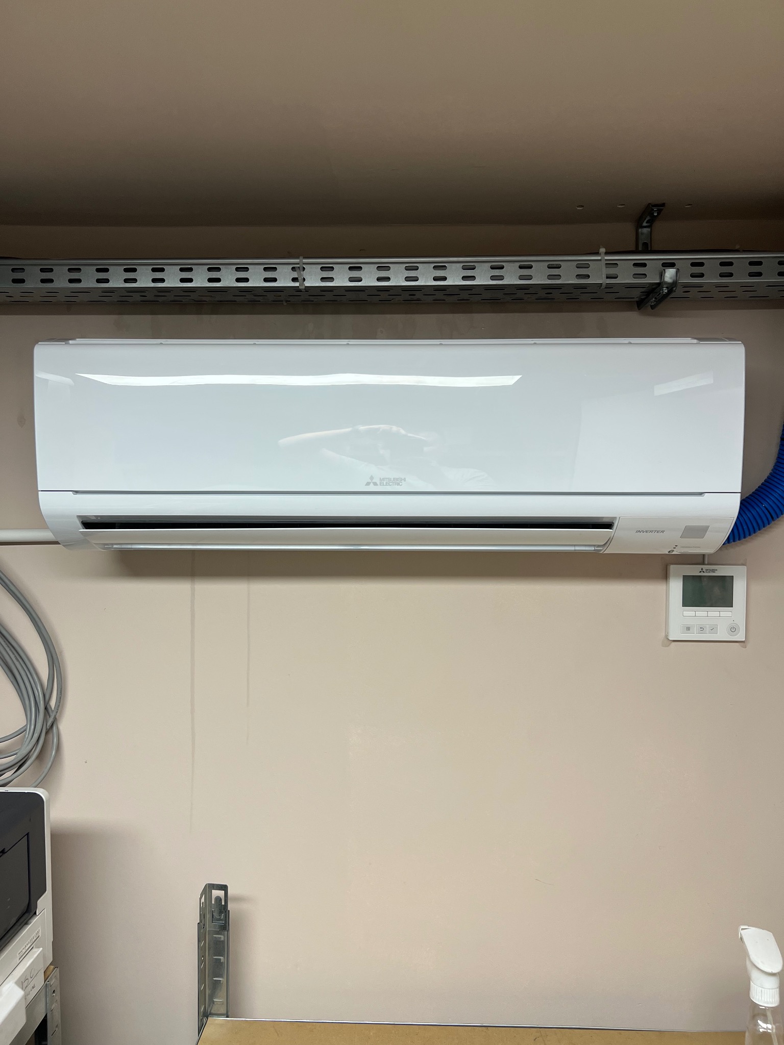 Biała klimatyzacja Mitsubishi Electric zamontowana na ścianie, widoczny sterownik i fragment niebieskiego przewodu wentylacyjnego. Nad urządzeniem metalowa listwa montażowa.