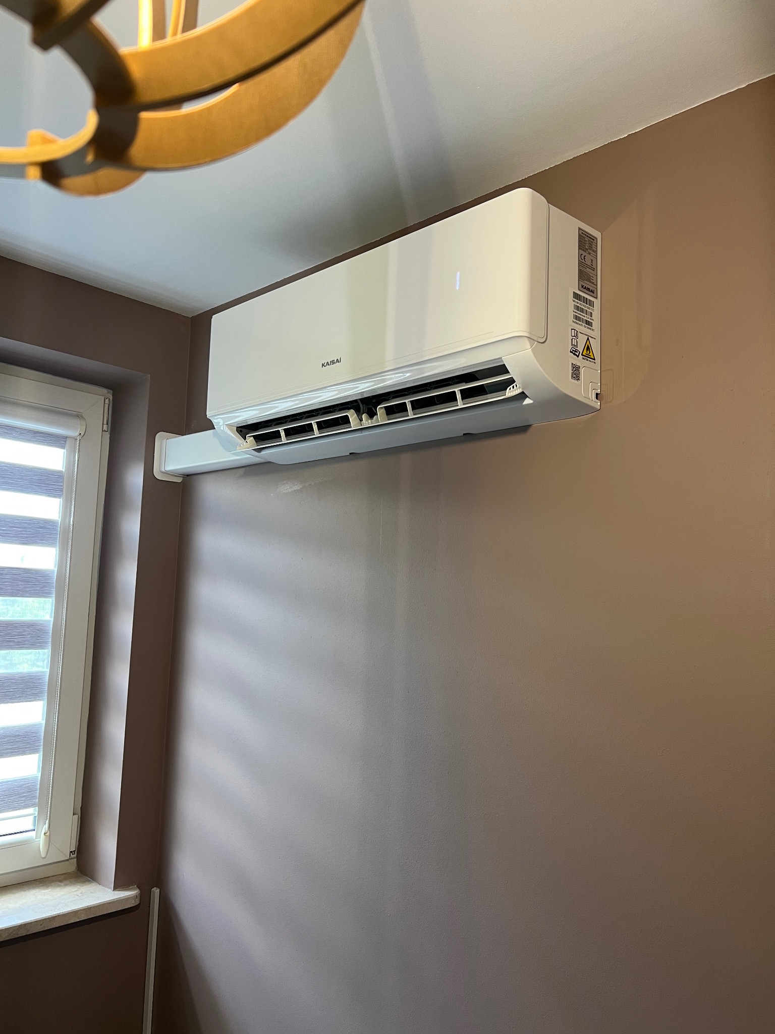 Biały klimatyzator Kaisi zamontowany na brązowej ścianie w pokoju. Widoczny fragment okna i lampy. Klimatyzacja w domu.