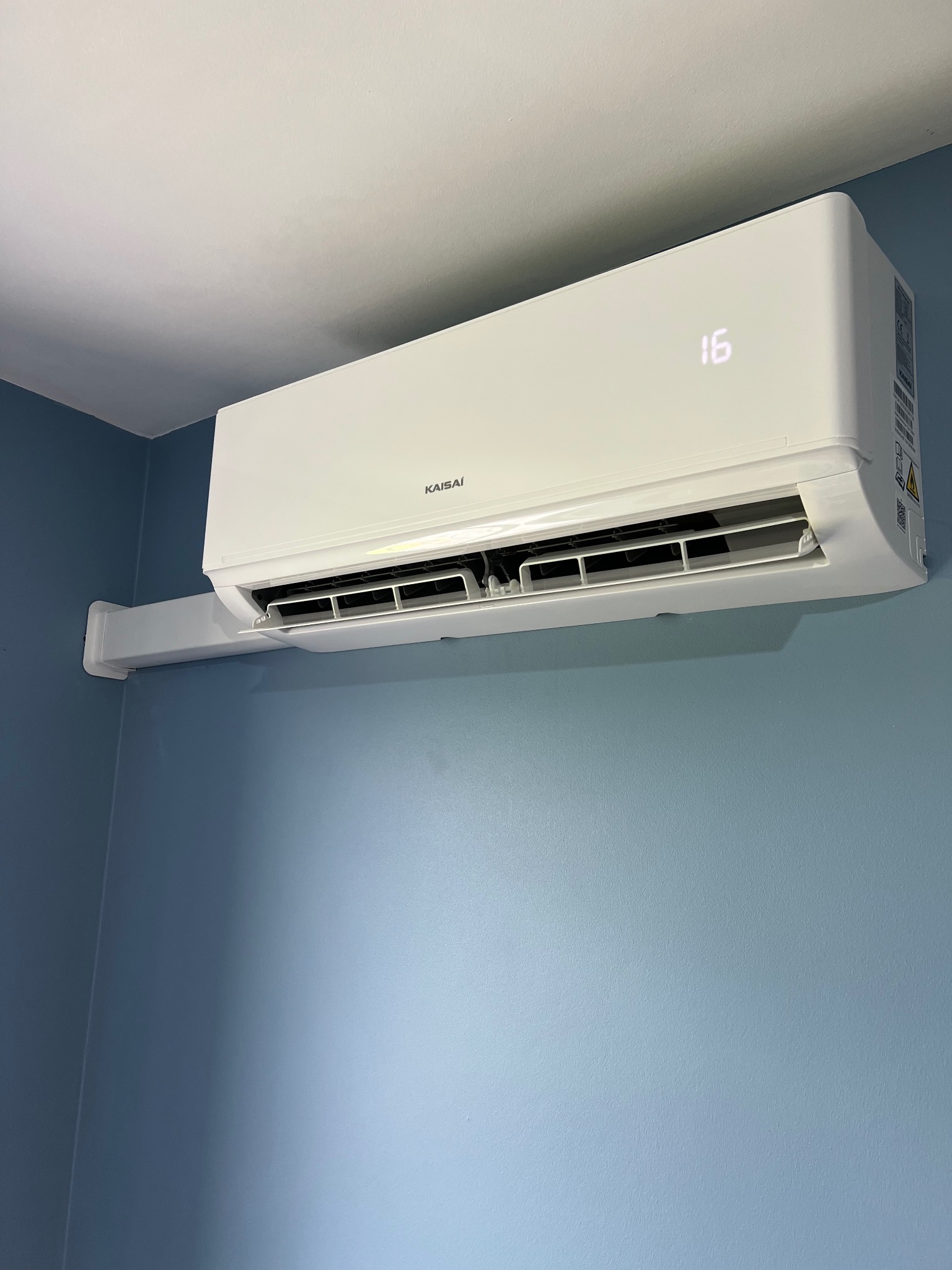 Nowoczesny, biały klimatyzator Kaisai z wyświetlaczem temperatury ustawiony na 16 stopni, zamontowany na błękitnej ścianie w pomieszczeniu.