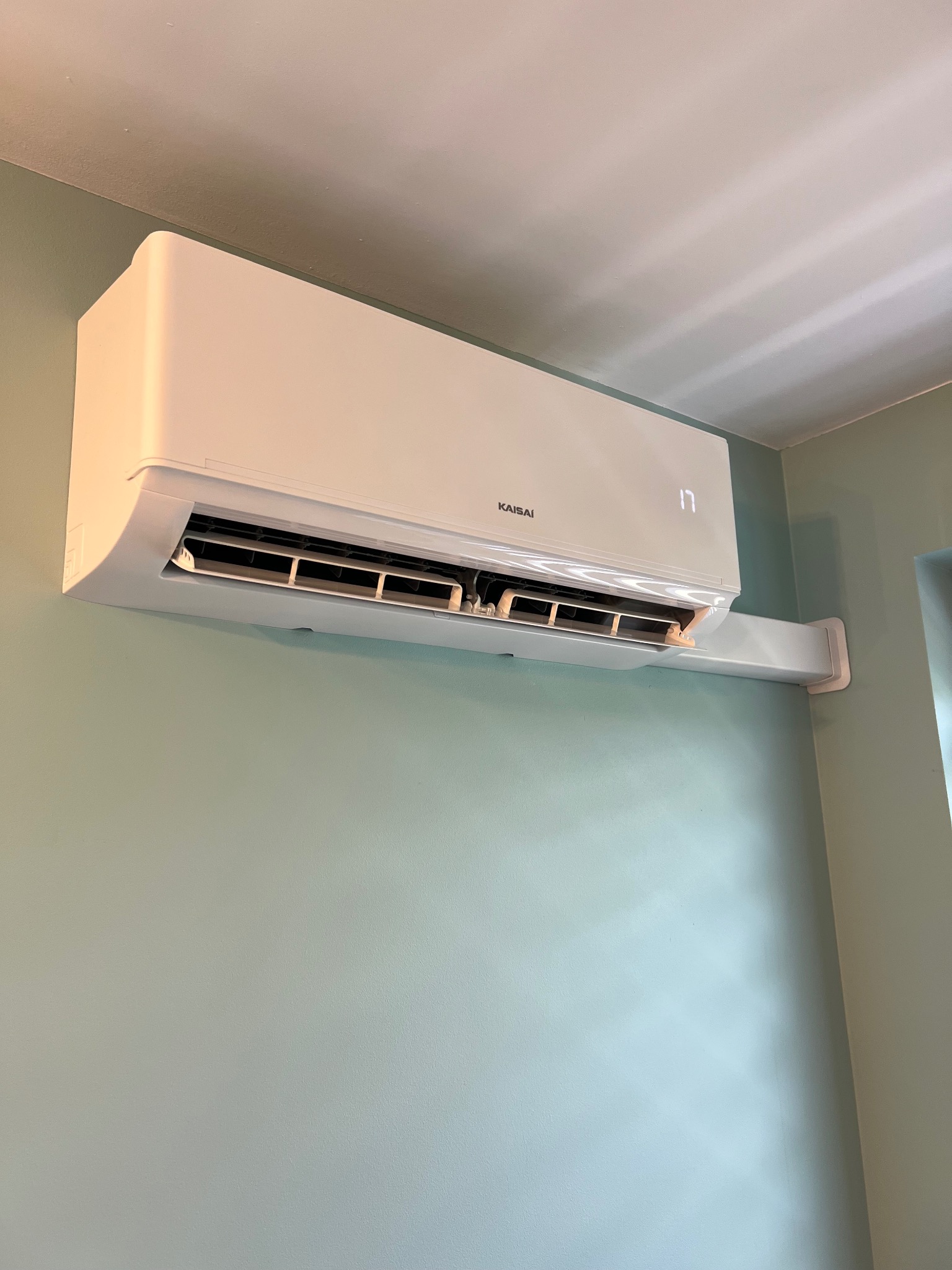 Biała klimatyzacja Kaisai z wyświetlaczem 17 stopni Celsjusza zamontowana na zielonej ścianie w rogu pokoju. Widoczne żaluzje i uchwyt montażowy.