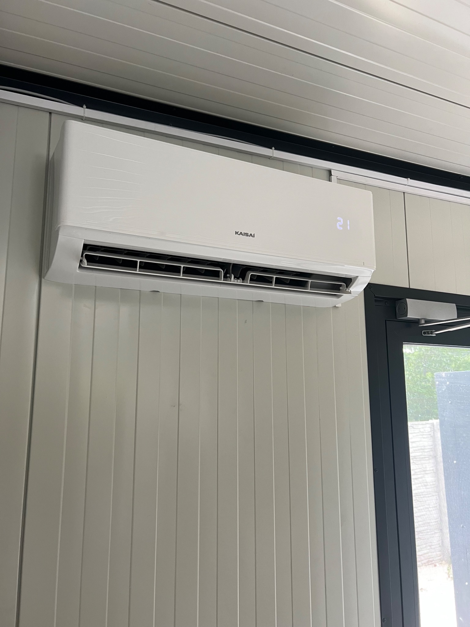 Biała klimatyzacja Kaisai zamontowana na ścianie z paneli, obok ciemne drzwi z samozamykaczem. Na wyświetlaczu widoczna temperatura 21 stopni.