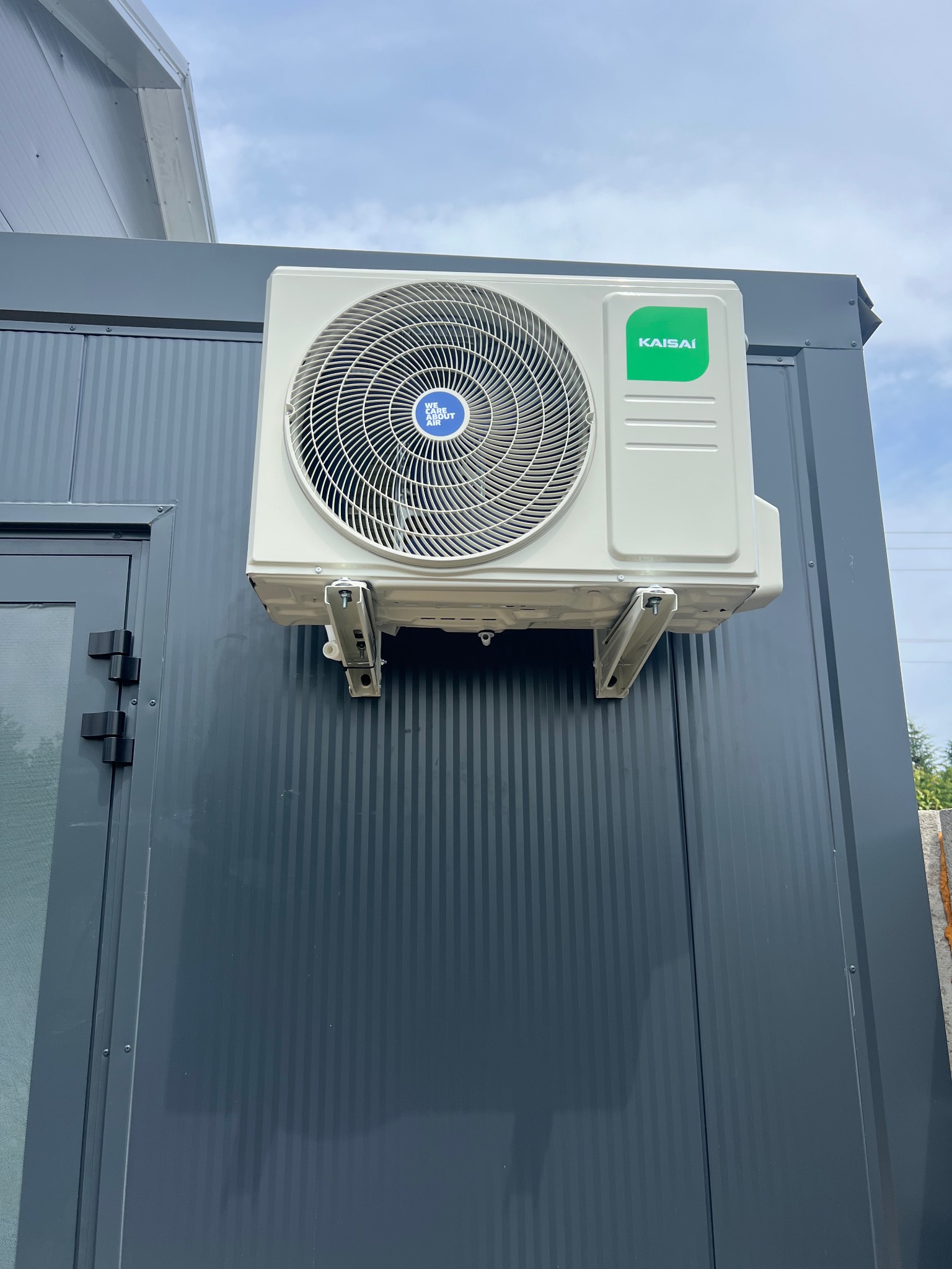 Biała klimatyzacja Kaisai zamontowana na szarej, pionowo żłobkowanej ścianie budynku, widok z dołu, lekko pochmurne niebo w tle.