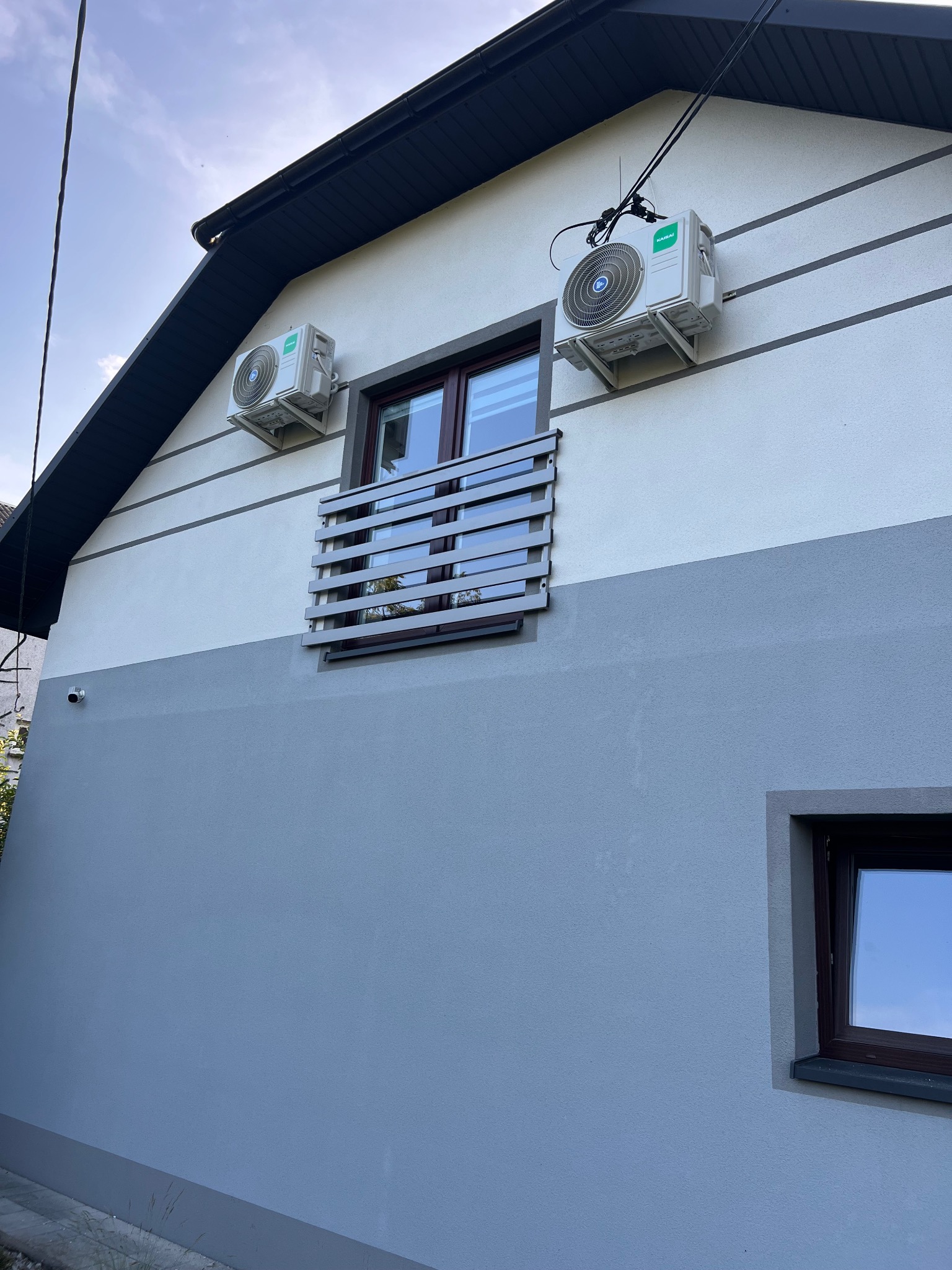 Elewacja domu z dwoma zamontowanymi klimatyzatorami marki Kaisai, umieszczonymi symetrycznie po obu stronach okna z balustradą. Widoczne przewody i instalacja.