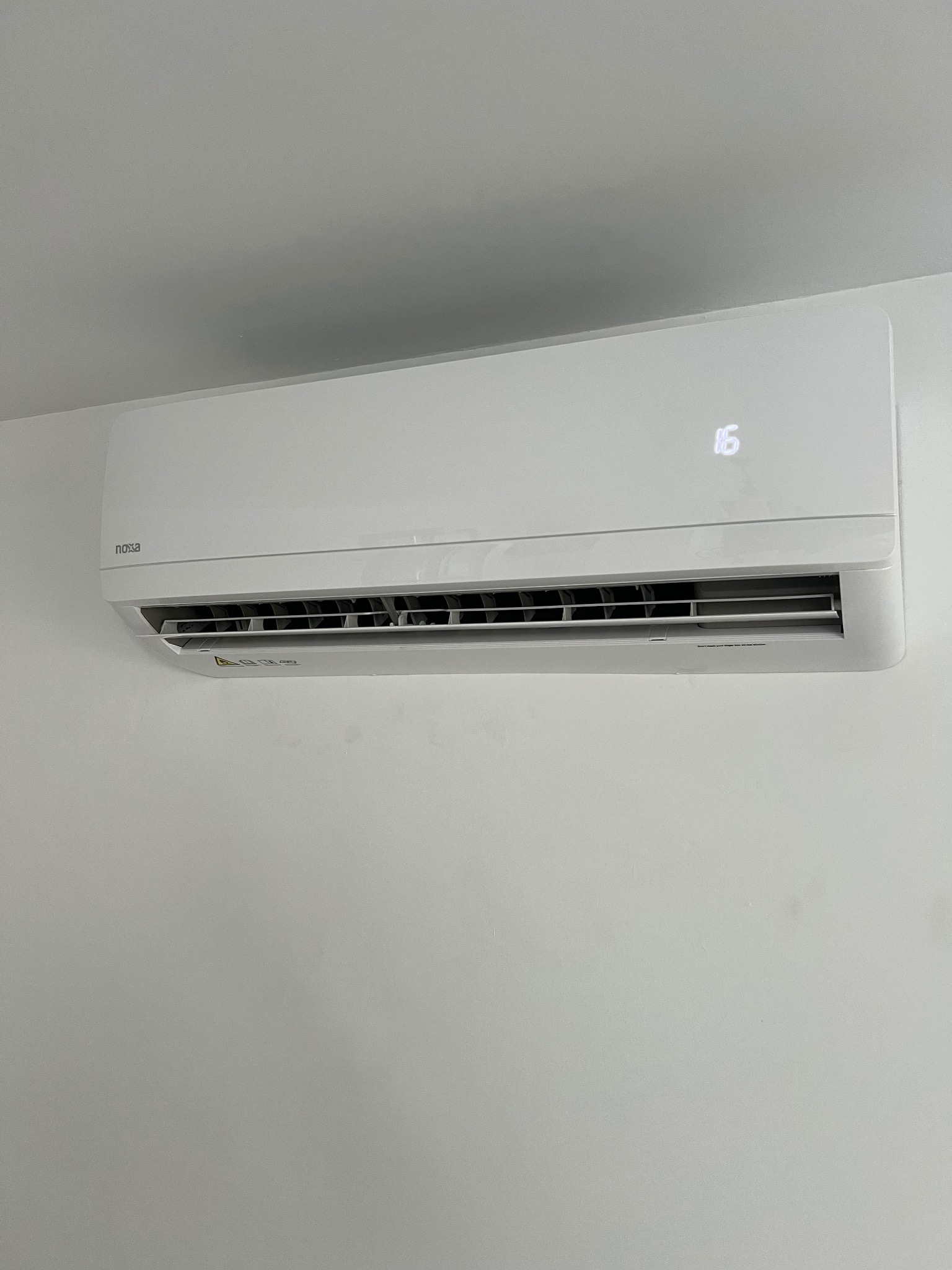 Biała klimatyzacja Nosa na jasnej ścianie. Widoczny wyświetlacz z temperaturą 16 stopni. Minimalistyczny design, czyste tło, wyraźne detale.