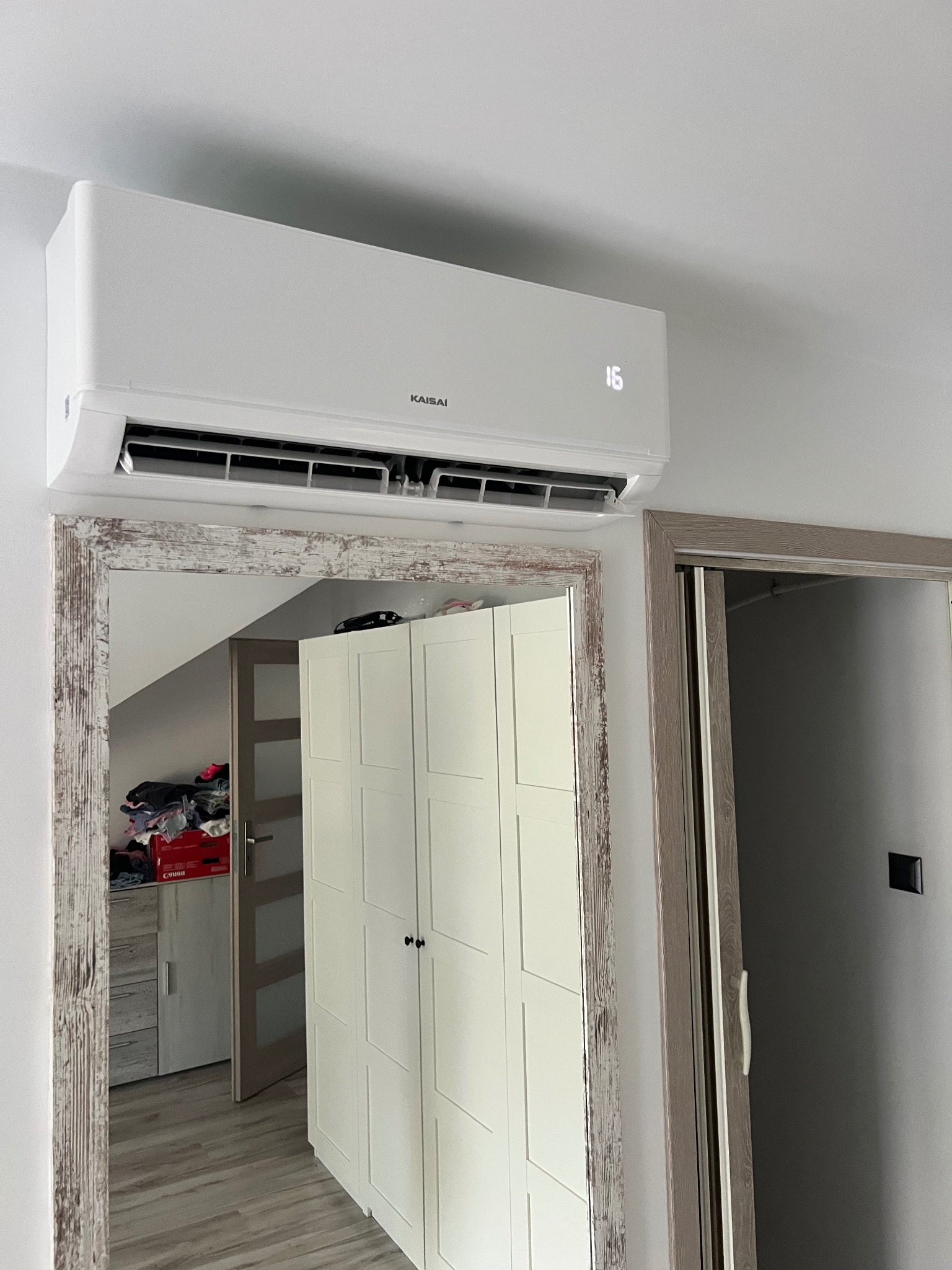 Biała klimatyzacja Kaisai zamontowana w pokoju, widoczny wyświetlacz z temperaturą 16 stopni. W tle lustro z drewnianą ramą i szafa.