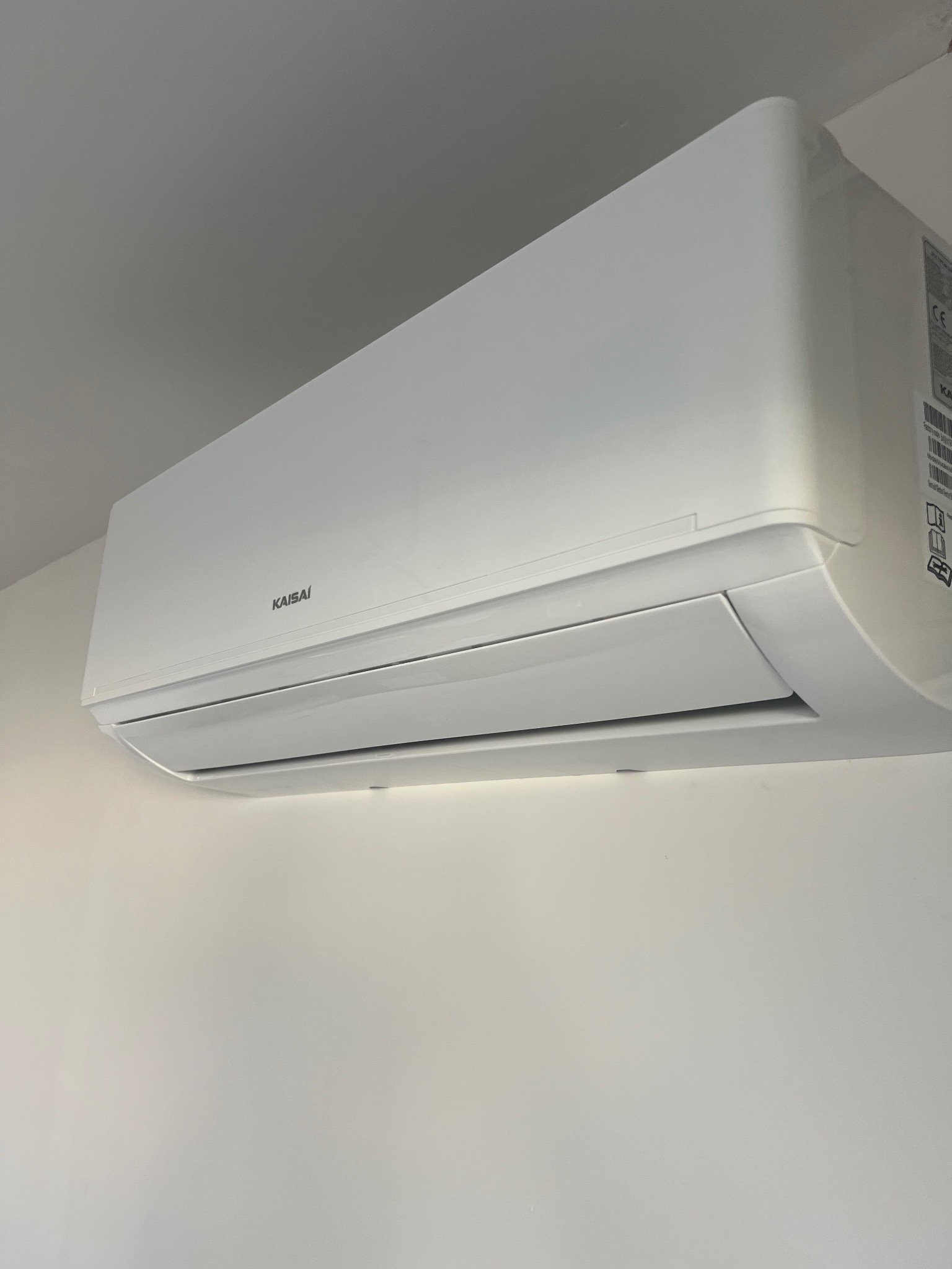 Biała klimatyzacja Kaisai zamontowana na jasnej ścianie, widoczna z boku, z elementami identyfikacyjnymi producenta. Minimalistyczny design.