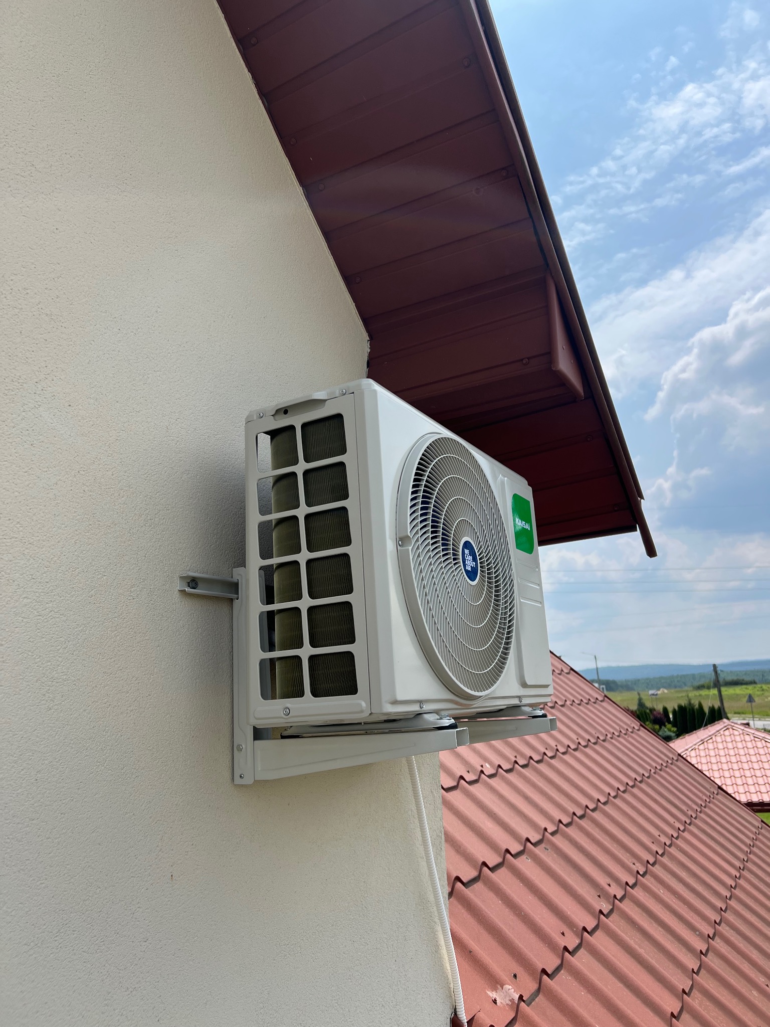 Biała jednostka zewnętrzna klimatyzacji Kaisai zamontowana na elewacji domu z widocznym dachem i fragmentem nieba w tle. Ujęcie z dołu.