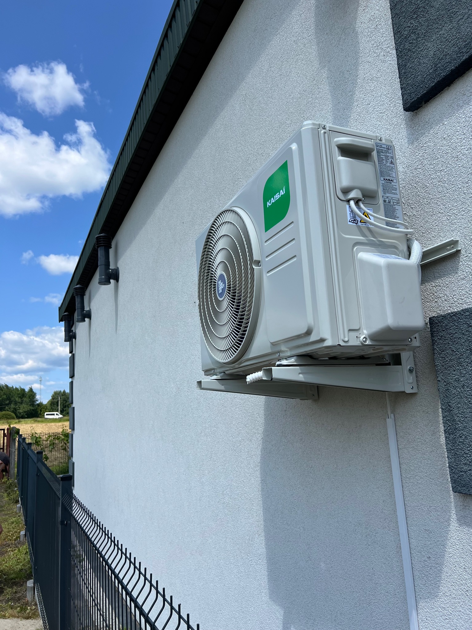Biały klimatyzator Kaisai zamontowany na elewacji budynku z szarą listwą, widok z dołu, ogrodzenie panelowe w tle, błękitne niebo.