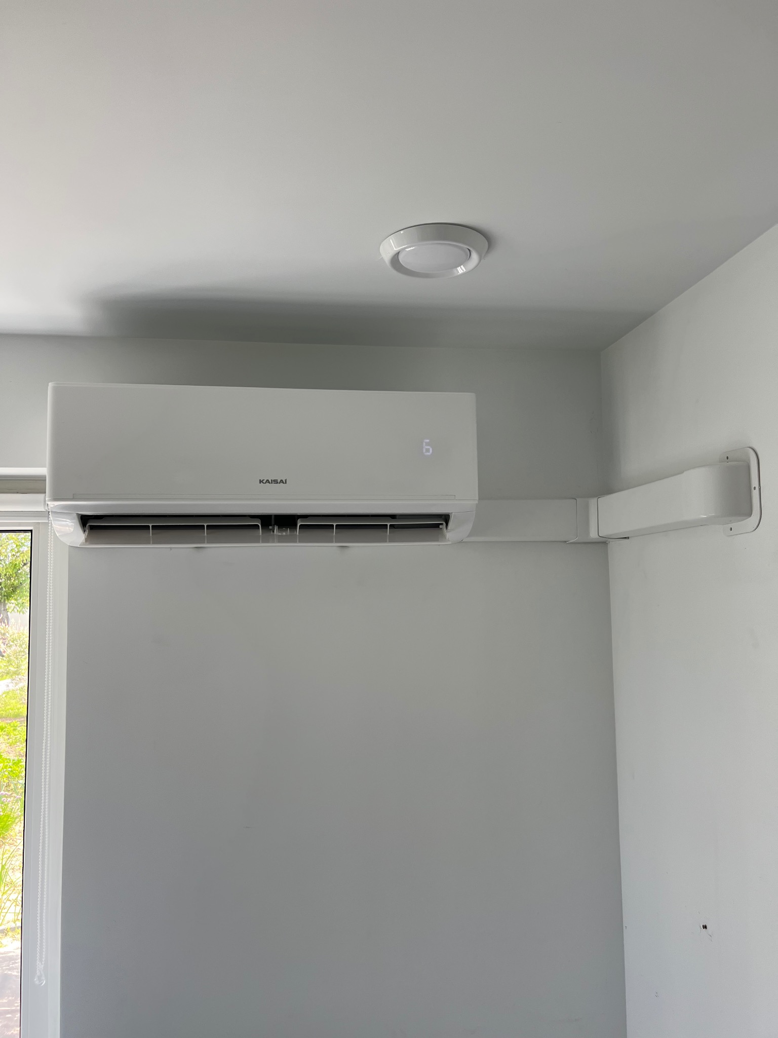 Biały klimatyzator Kaisai zamontowany na ścianie w jasnym pomieszczeniu. Widoczna listwa maskująca instalację elektryczną i chłodniczą. Minimalistyczny design.