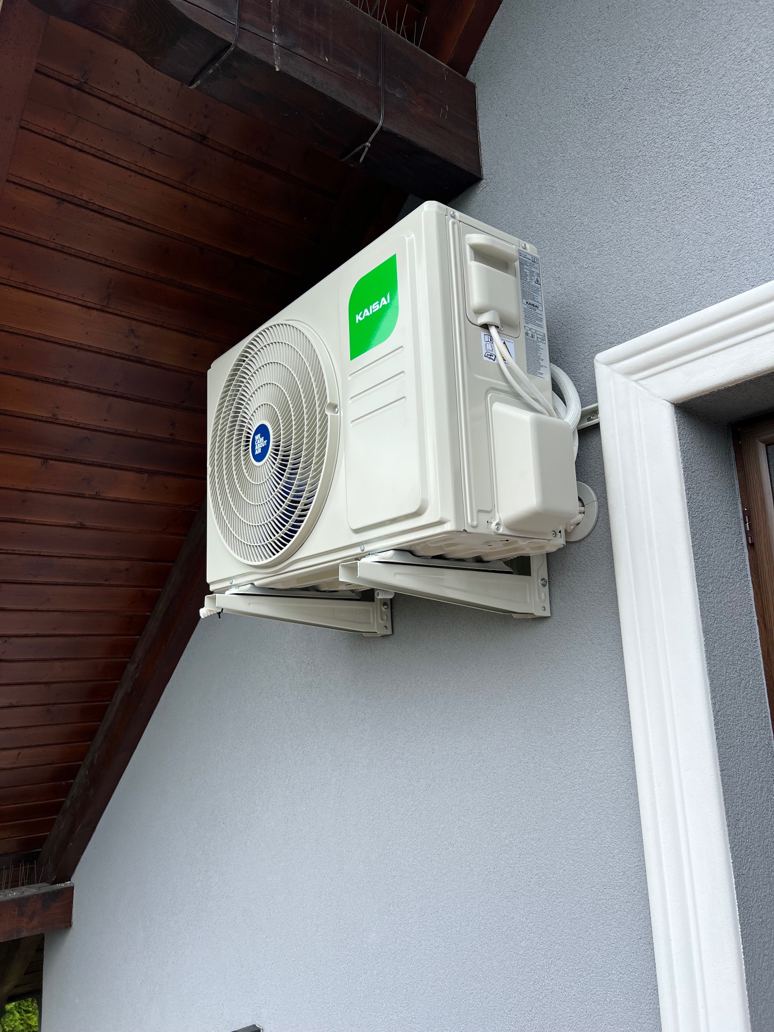 Zewnętrzna jednostka klimatyzacji Kaisai zamontowana na szarej ścianie budynku z drewnianym okapem. Widoczne białe rurki i uchwyty montażowe.