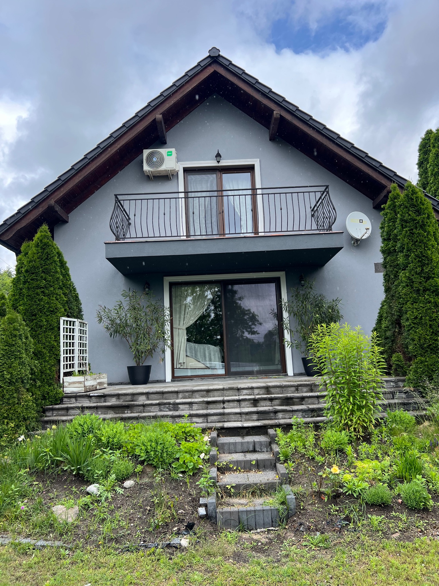 Szary dom z balkonem i klimatyzatorem Haier na ścianie. Widoczne rośliny w donicach i satelita. Dom otoczony zielenią w pochmurny dzień.