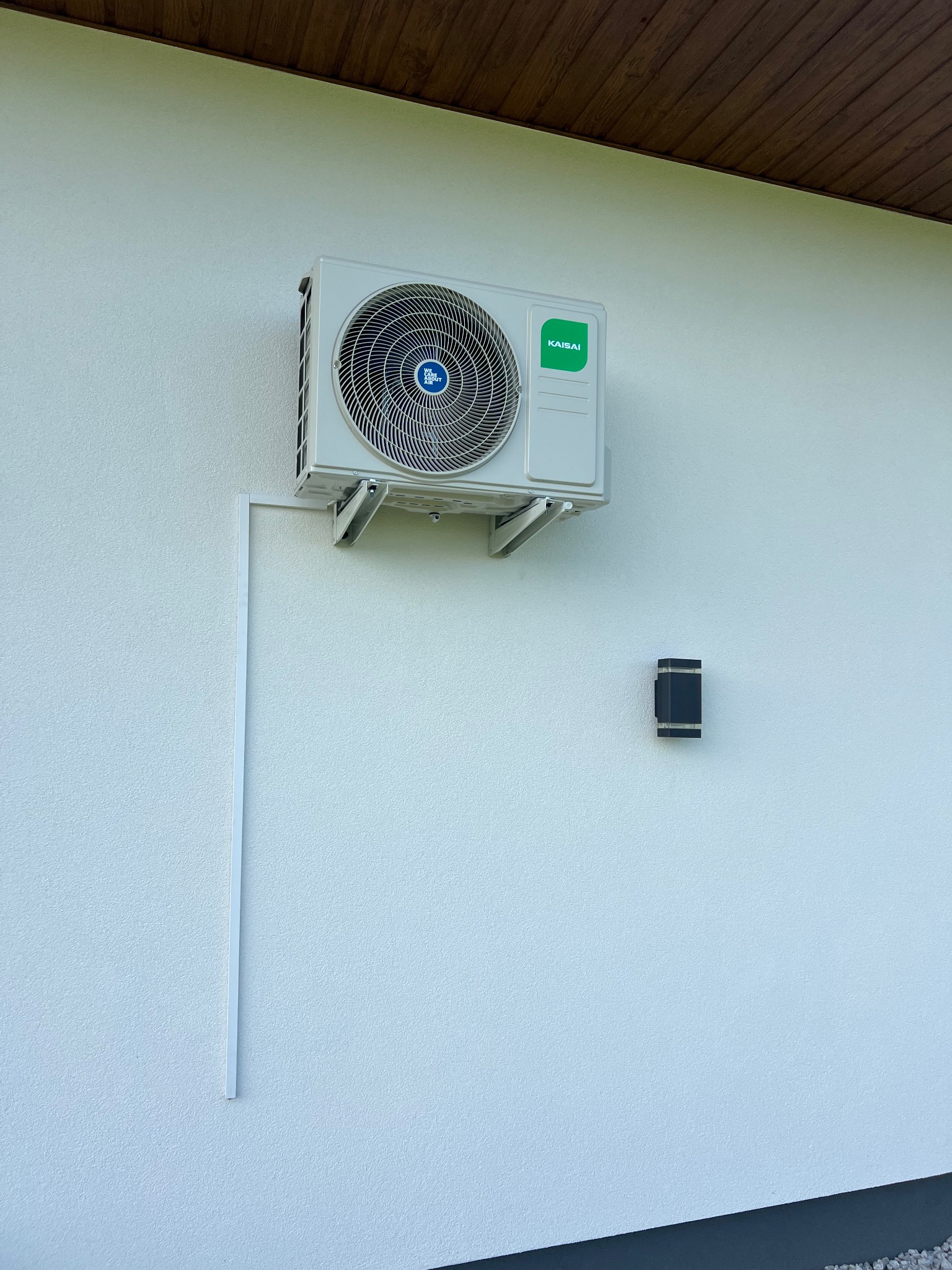 Klimatyzator Kaisai zamontowany na białej elewacji budynku, widoczne rurki instalacyjne i lampa ścienna. Estetyczny montaż.