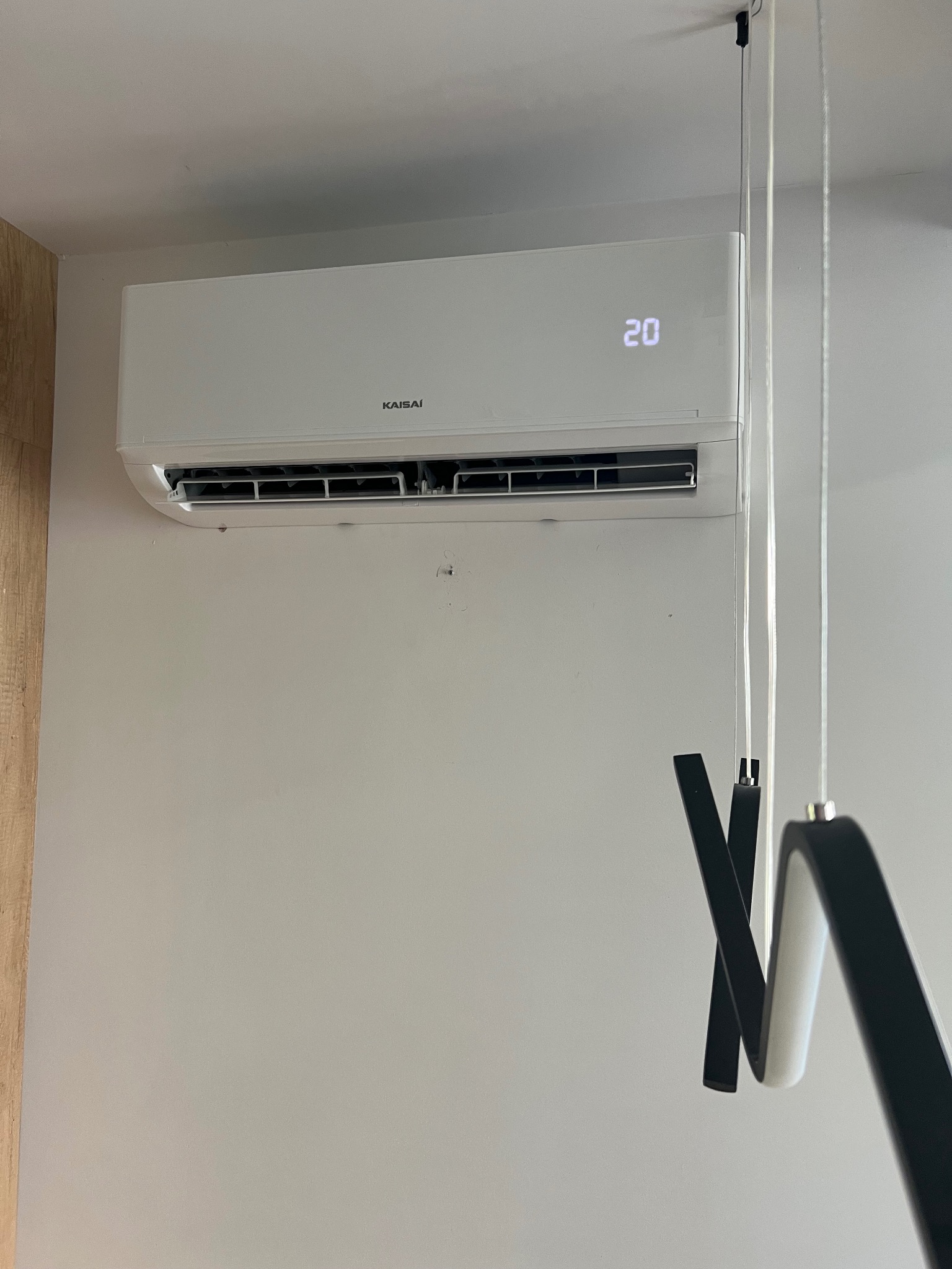 Biała klimatyzacja Kaisai na ścianie, widoczny wyświetlacz z temperaturą 20 stopni Celsjusza, obok nowoczesna lampa wisząca.