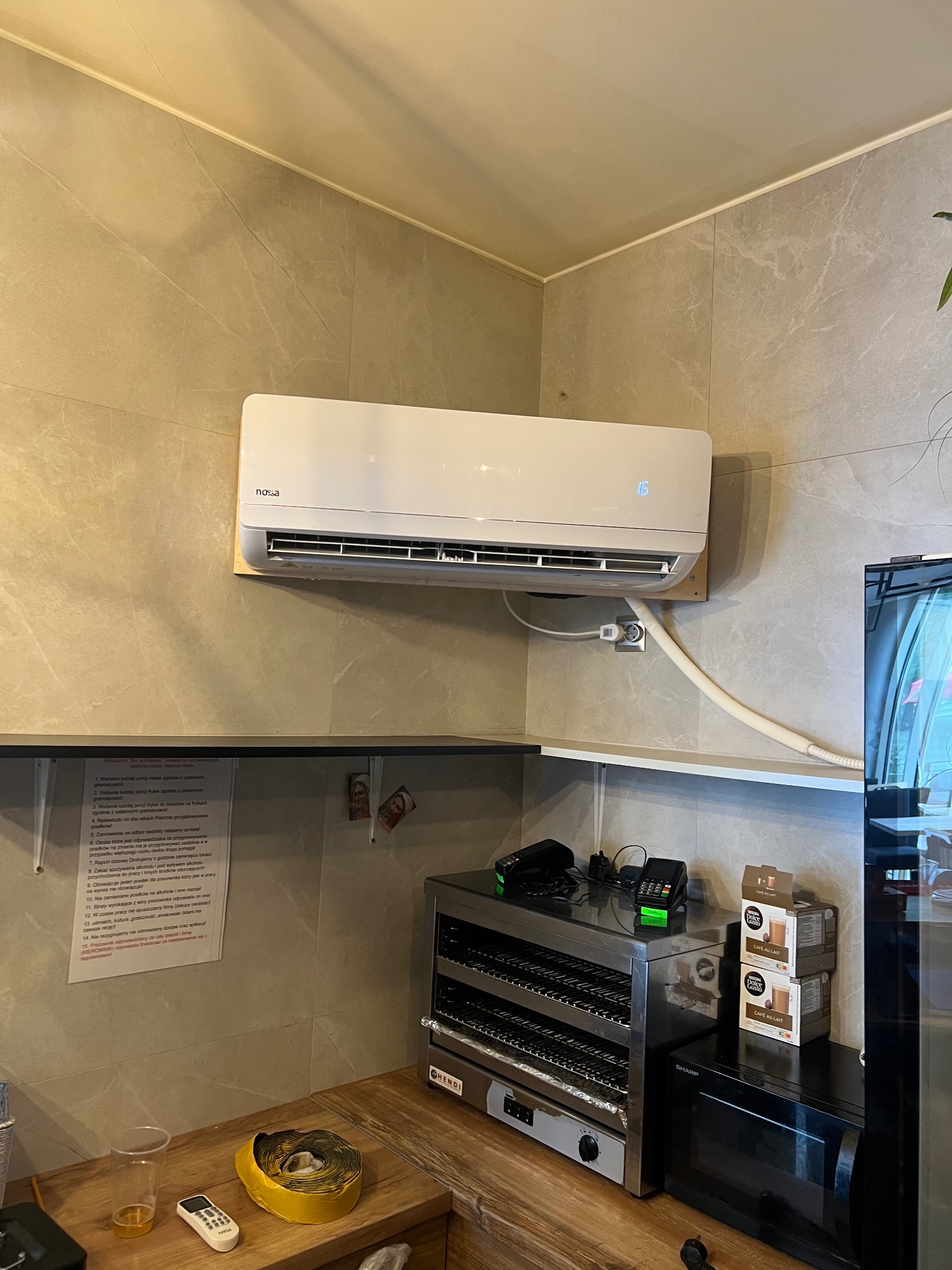 Klimatyzator NOXA zamontowany na ścianie z szarych płytek imitujących marmur w lokalu gastronomicznym. Widoczne półki, urządzenia kuchenne i instrukcja obsługi.