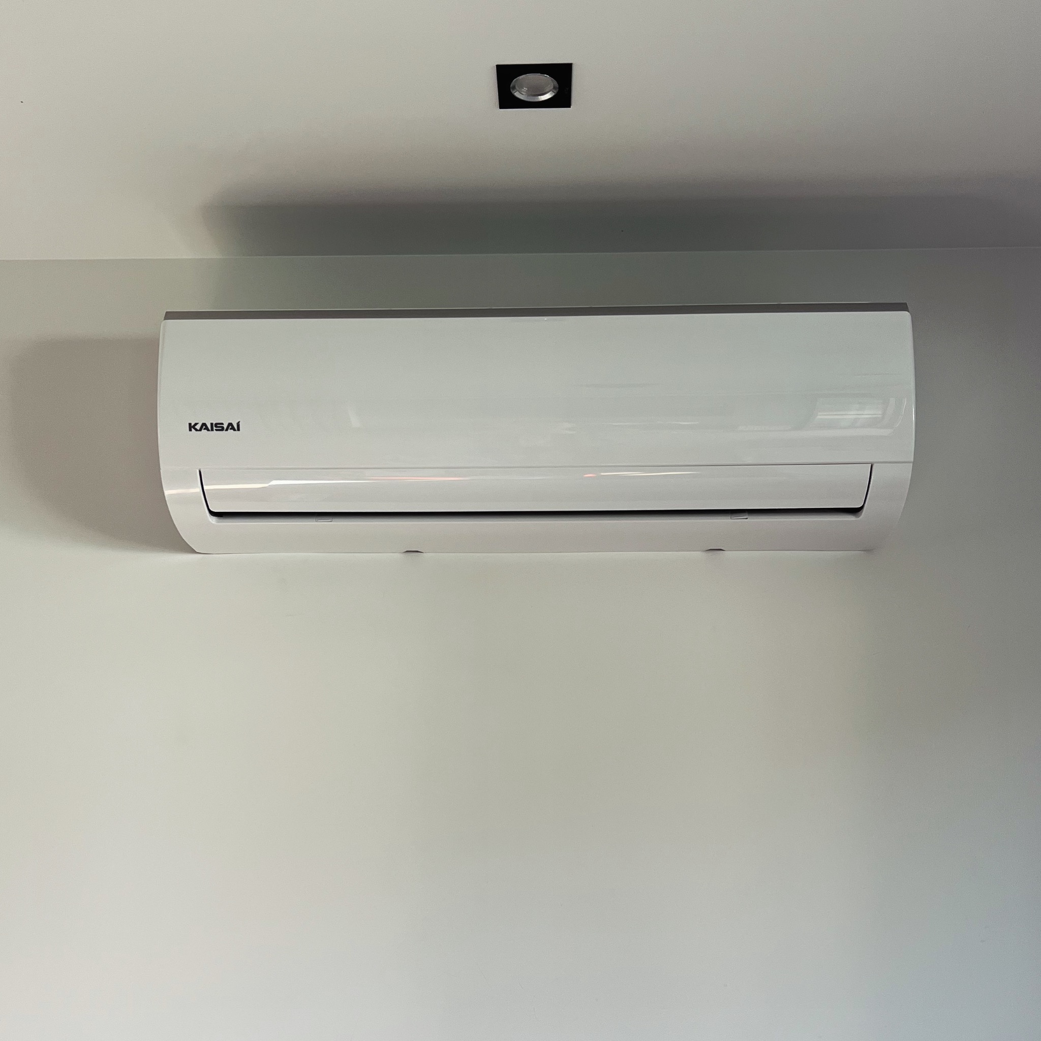 Biały klimatyzator Kaisai zamontowany na jasnej ścianie, pod kwadratową lampą sufitową. Minimalistyczny design, widoczne detale urządzenia.