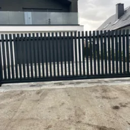 Nowoczesna, przesuwna brama w kolorze antracytowym z pionowymi profilami, zamontowana przy posesji z białym domem i szarą dachówką, widok z zewnątrz na posesję.