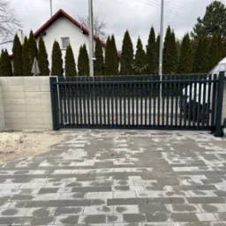 Nowoczesna, przesuwna brama w kolorze grafitowym z automatyką, zamontowana przy betonowym ogrodzeniu modułowym i wybrukowanym podjeździe. W tle rząd wysokich, zielonych tuj.