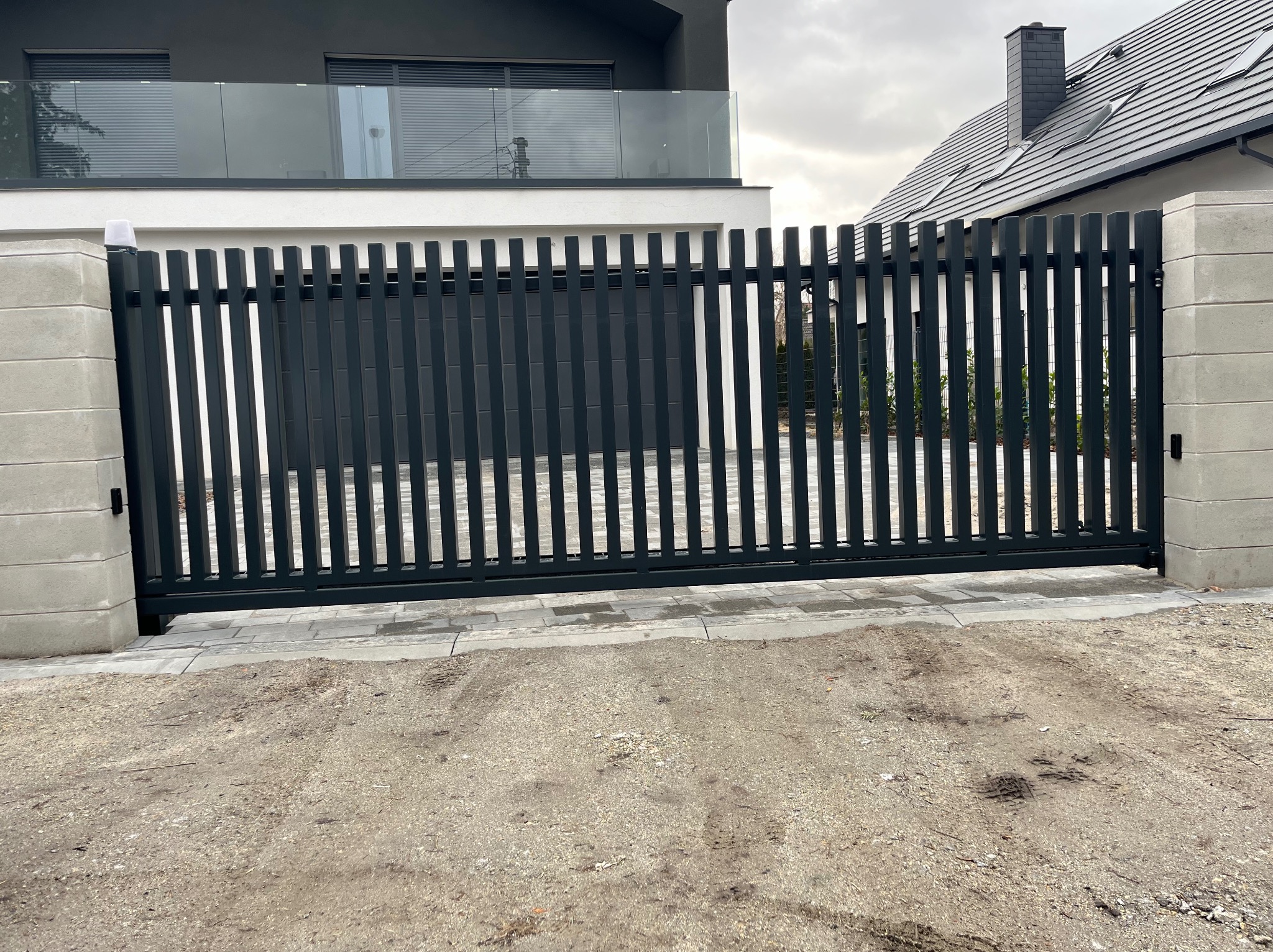 Nowoczesna, przesuwna brama w kolorze antracytowym z pionowymi profilami, zamontowana przy posesji z białym domem i szarą dachówką, widok z zewnątrz na posesję.