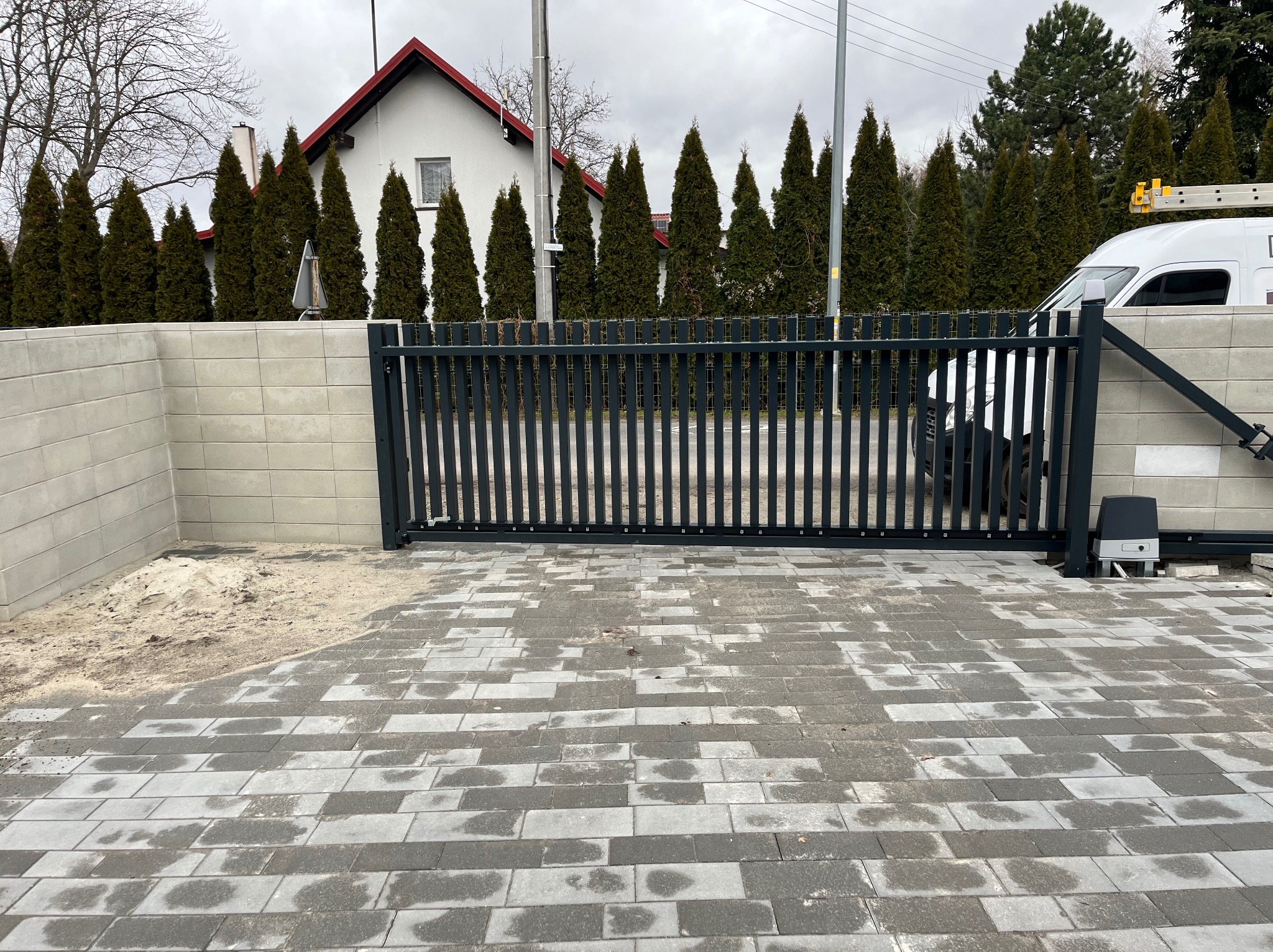 Nowoczesna, przesuwna brama w kolorze grafitowym z automatyką, zamontowana przy betonowym ogrodzeniu modułowym i wybrukowanym podjeździe. W tle rząd wysokich, zielonych tuj.