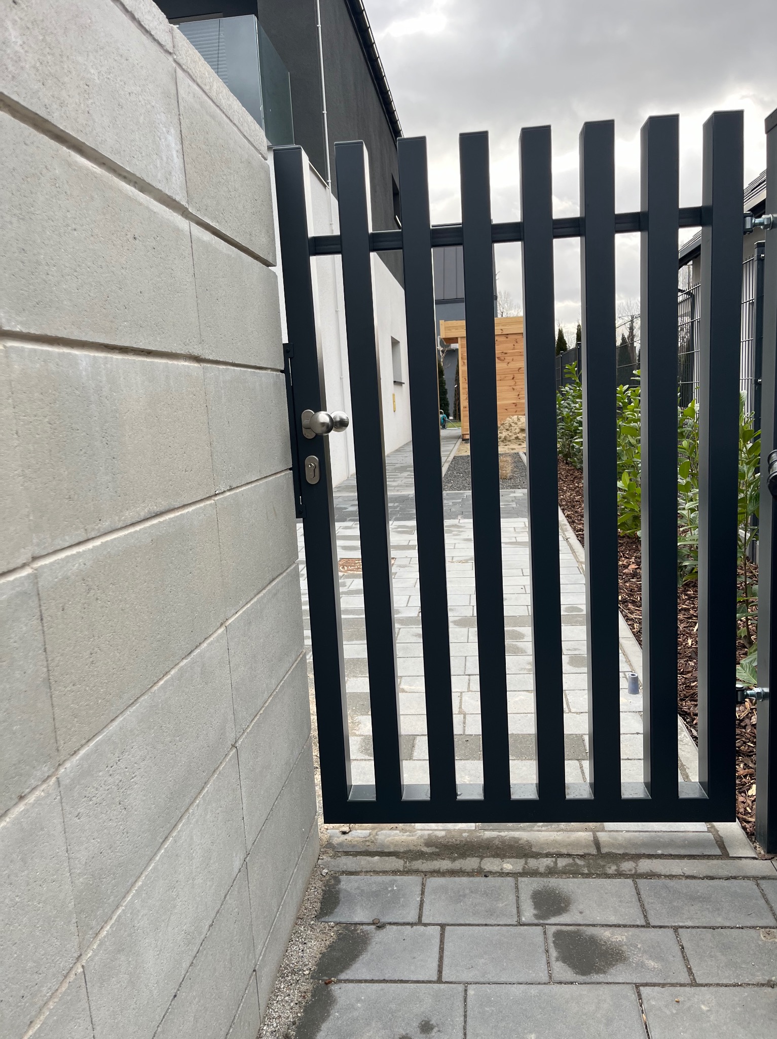 Otwarta, nowoczesna brama w kolorze antracytowym, z pionowymi profilami, zamontowana przy murze z betonowych bloczków. Widoczny fragment posesji z wybrukowaną ścieżką i roślinnością.