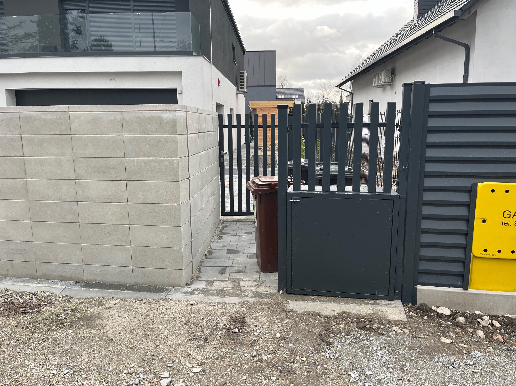 Nowoczesne, grafitowe ogrodzenie z furtką i bramą wjazdową, widok z zewnątrz posesji na tle jasnych budynków mieszkalnych i kostki brukowej.