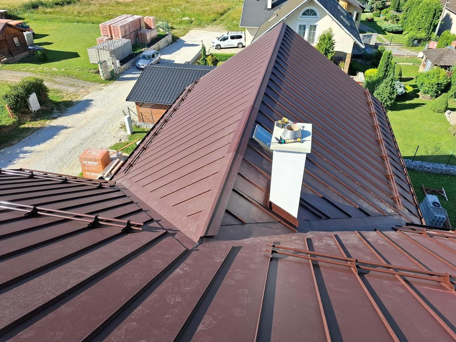 Widok z góry na brązowy dach z blachy trapezowej z kominem i akcesoriami montażowymi, w tle domy i zielona trawa.