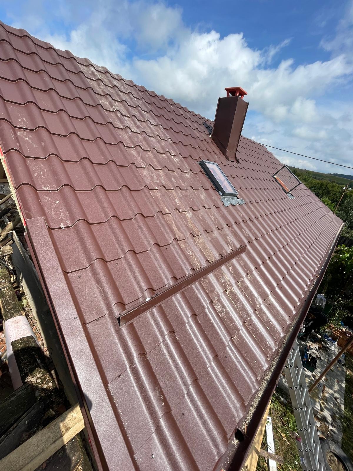 Ciemnobrązowy dach z blachy trapezowej, widoczne dwa okna dachowe i komin, tło to błękitne niebo i zieleń drzew