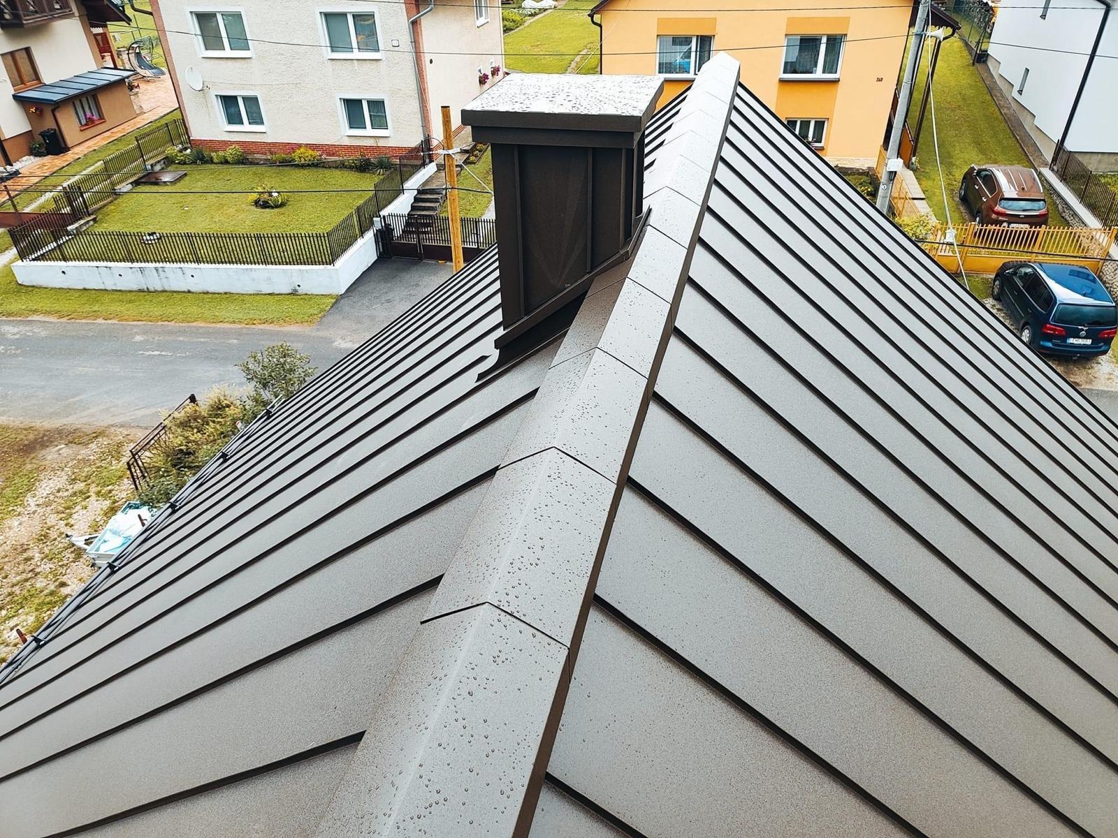 Nowoczesny, brązowy dach z blachy trapezowej z kominem, widok z góry, krople deszczu na powierzchni, w tle zabudowa jednorodzinna i zieleń.