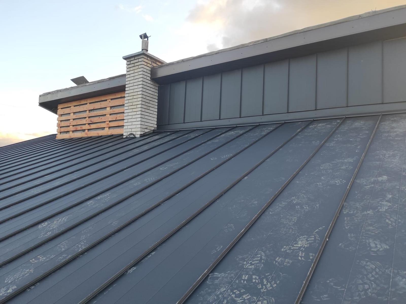 Metalowy dach w kolorze grafitowym z widocznymi śladami stóp, komin z jasnej cegły oraz drewniana konstrukcja osłaniająca element dachu.