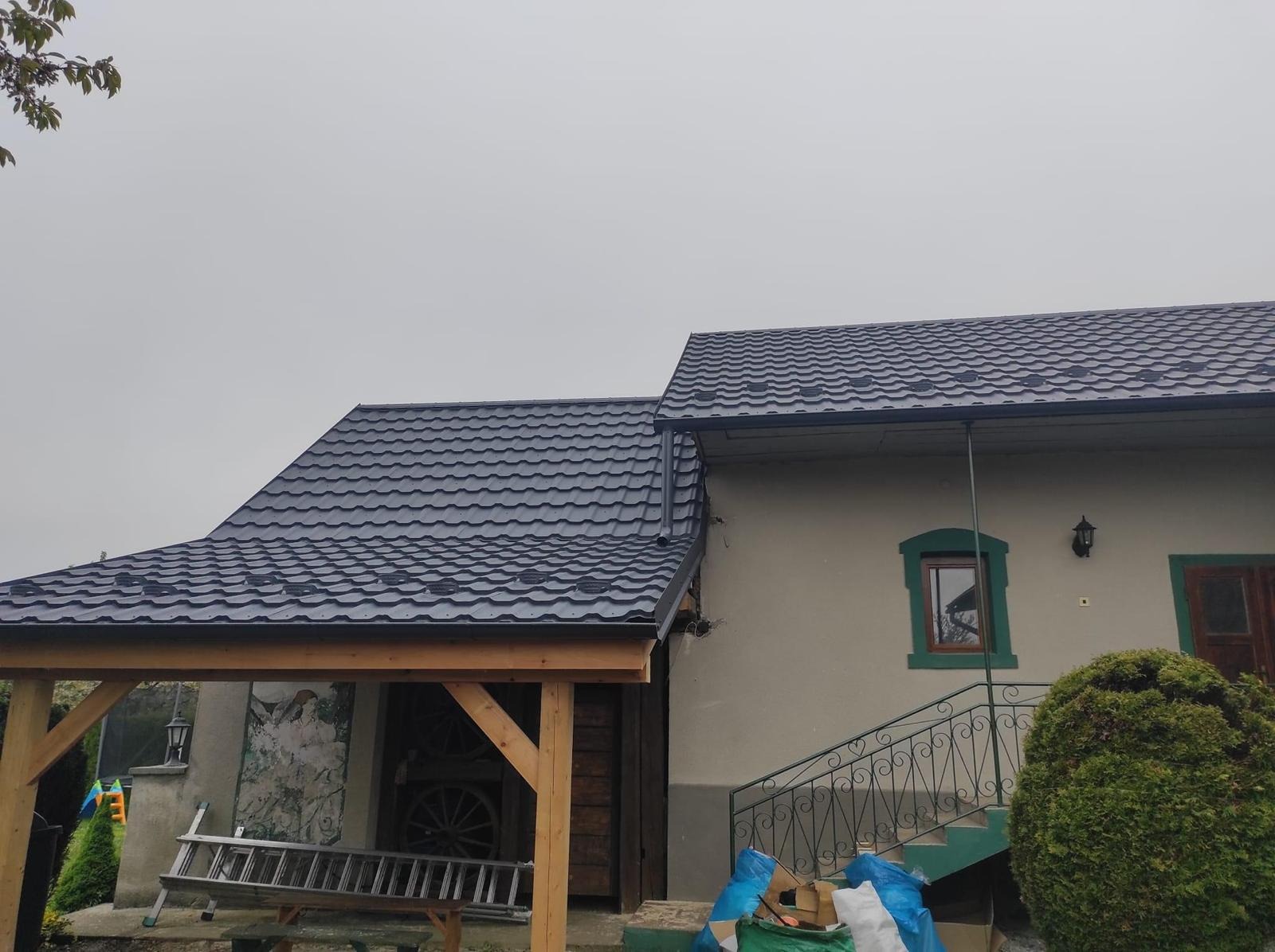 Dom jednorodzinny z nowym, ciemnoszarym dachem z blachodachówki, widoczne rynny i drewniana wiata z boku, drabina oparta o ścianę, pochmurne niebo.