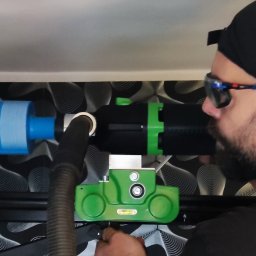 TermoService - Mężczyzna w czapce i okularach, z profilu, używa specjalistycznego sprzętu do pomiaru instalacji wentylacyjnej z niebieskim kanałem, na tle ściany z wzorem.