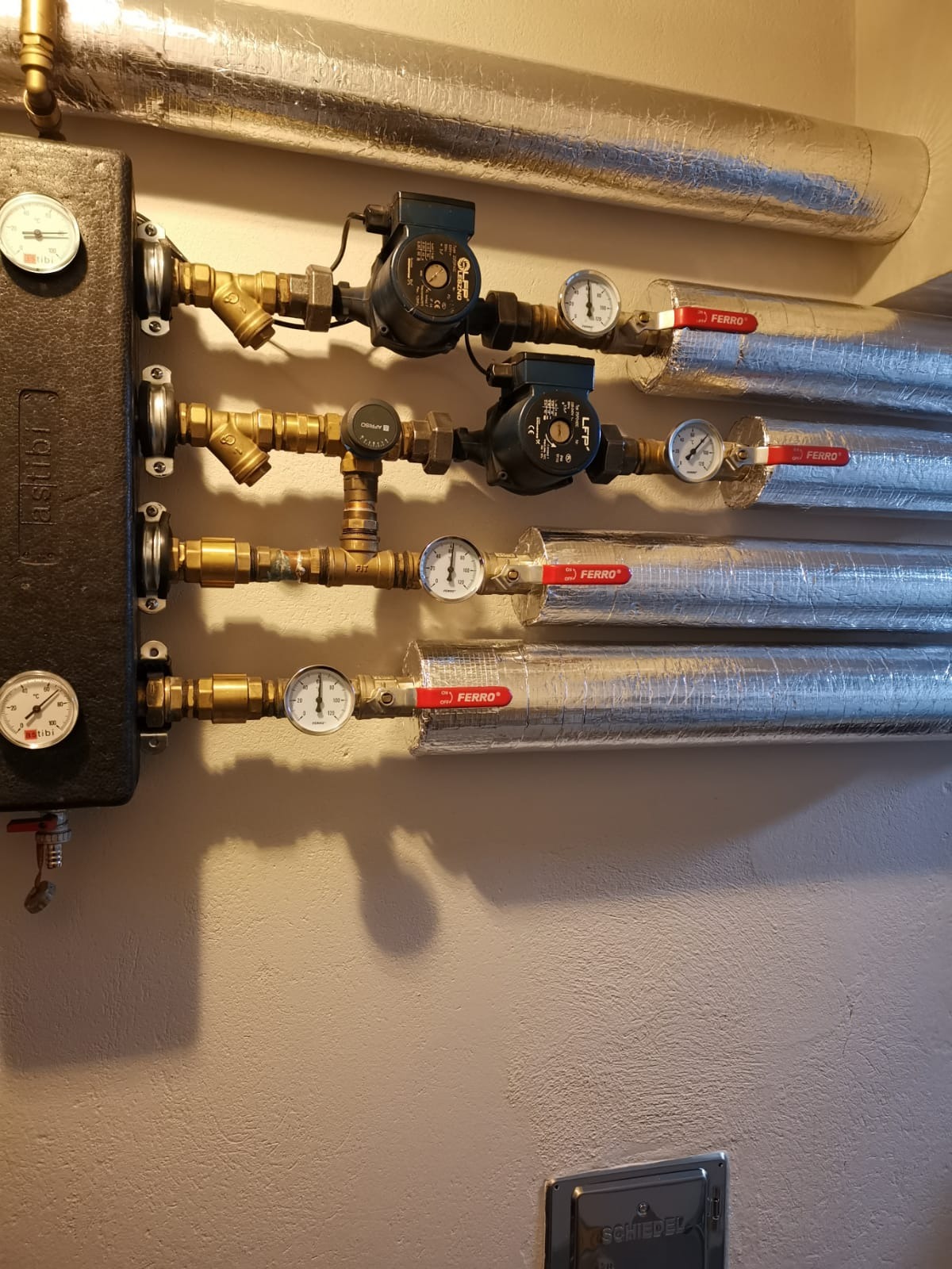 Fragment instalacji centralnego ogrzewania z widocznymi rurami w srebrnej izolacji, zaworami Ferro, manometrami, pompami obiegowymi i czarnym panelem sterującym Astibi.