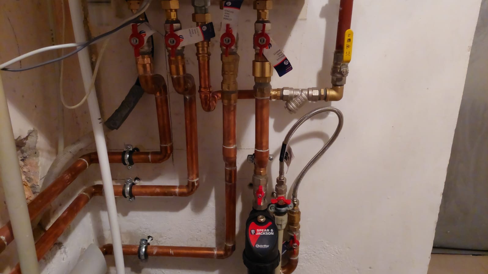 Miedziane rury instalacji gazowej i wodnej z zaworami odcinającymi, filtrem magnetycznym Spear & Jackson BoilerMag i elastycznym przewodem przyłączeniowym na tle ściany.