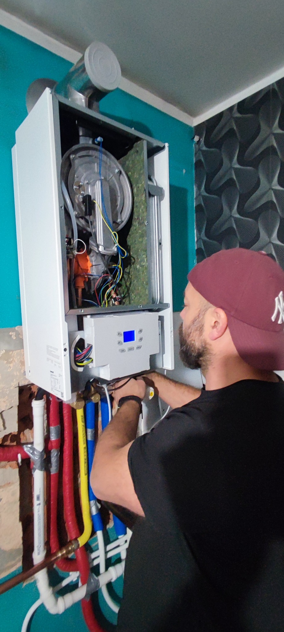 Mężczyzna w czarnej koszulce i bordowej czapce z daszkiem podłącza przewody do otwartego pieca gazowego wiszącego na ścianie z widocznymi rurami instalacji grzewczej w kolorach czerwonym, żółtym...