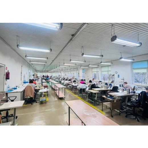 Wnętrze hali produkcyjnej z rzędami stanowisk do szycia, oświetlonych lampami jarzeniowymi, z widocznymi pracownikami przy maszynach i półkami na materiały.