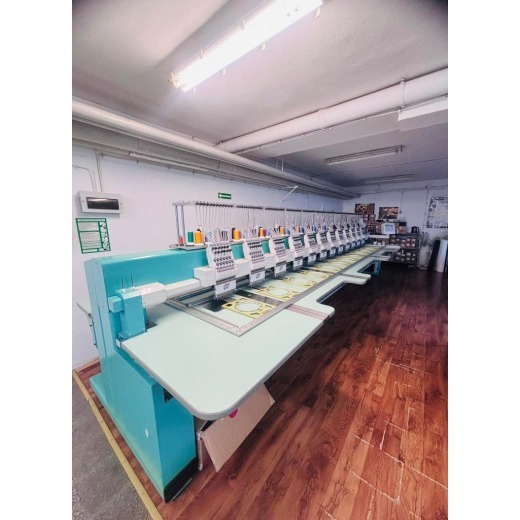 Długi rząd maszyn do haftu komputerowego w hali produkcyjnej, widoczne nici w różnych kolorach i wzory w trakcie tworzenia.