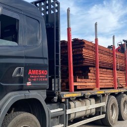 Tartak Amesko Spółka Jawna Monika Skóra-Szydłowska,Artur Szydłowski - Ciężarówka z czarną kabiną marki Scania, załadowana równo ułożonymi deskami z drewna, zabezpieczonymi czerwonymi słupkami, widoczne logo firmy transportowej AMESKO