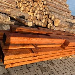 Tartak Amesko Spółka Jawna Monika Skóra-Szydłowska,Artur Szydłowski - Stos belek z obrobionego drewna na palecie, w tle pryzma okrąglaków i kostka brukowa.