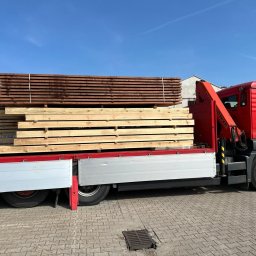 Tartak Amesko Spółka Jawna Monika Skóra-Szydłowska,Artur Szydłowski - Czerwona ciężarówka z dźwigiem Palfinger przewozi dwa stosy drewna: jasne deski na dole i ciemniejsze, impregnowane na górze, zabezpieczone białym sznurkiem.