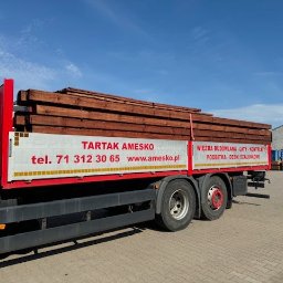 Tartak Amesko Spółka Jawna Monika Skóra-Szydłowska,Artur Szydłowski - Ciężarówka z czerwonym nadwoziem, załadowana brązową tarcicą budowlaną, z widocznym logo tartaku Amesko i numerem telefonu na boku, na tle błękitnego nieba i hali magazynowej.