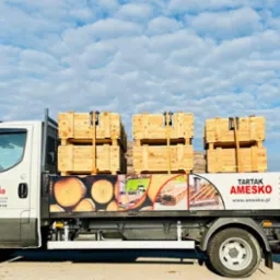 Ciężarówka z logo Tartak Amesko przewozi skrzynie z drewnem, widoczne także grafiki z przekrojami drewna i listą usług: więźba dachowa, impregnacja drewna, łaty, deska szalunkowa, podbitka dachowa,...