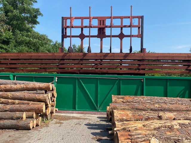 Stosy okrąglaków ułożone na placu przed zieloną bramą, za którą widoczna jest konstrukcja tartaku i brązowe ogrodzenie z desek.