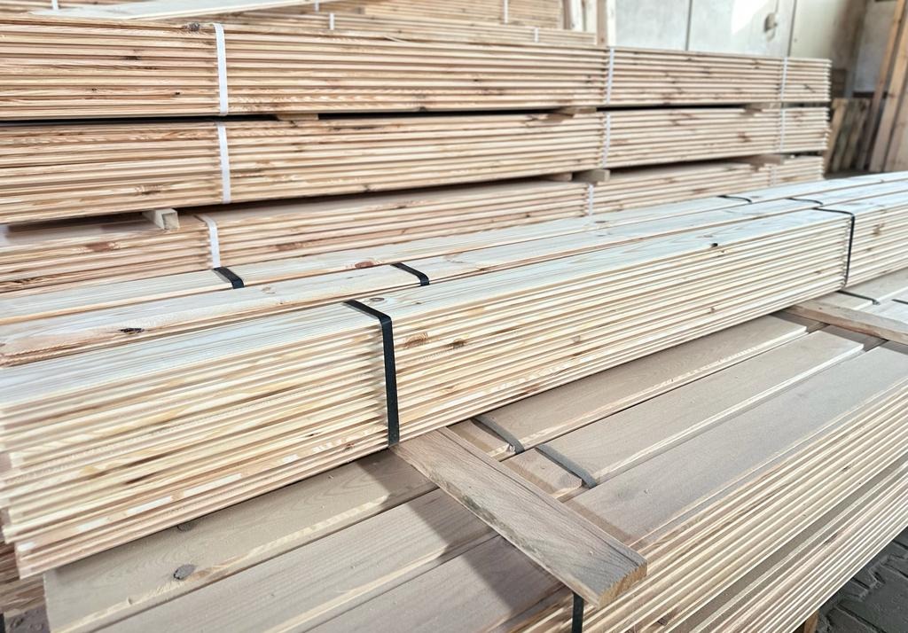 Stosy świeżo przetartej, jasnej tarcicy iglastej, spięte czarną taśmą, ułożone w hali.