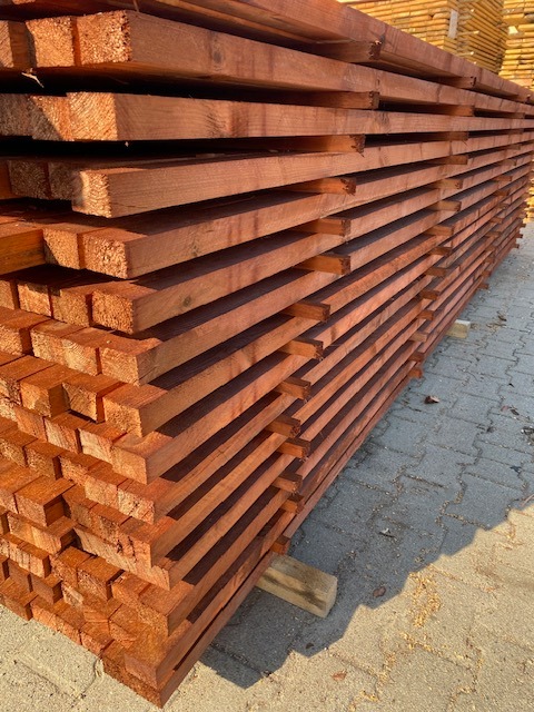 Stos czerwonej tarcicy budowlanej ułożonej na kostce brukowej, widoczne słoje drewna i ostre krawędzie.